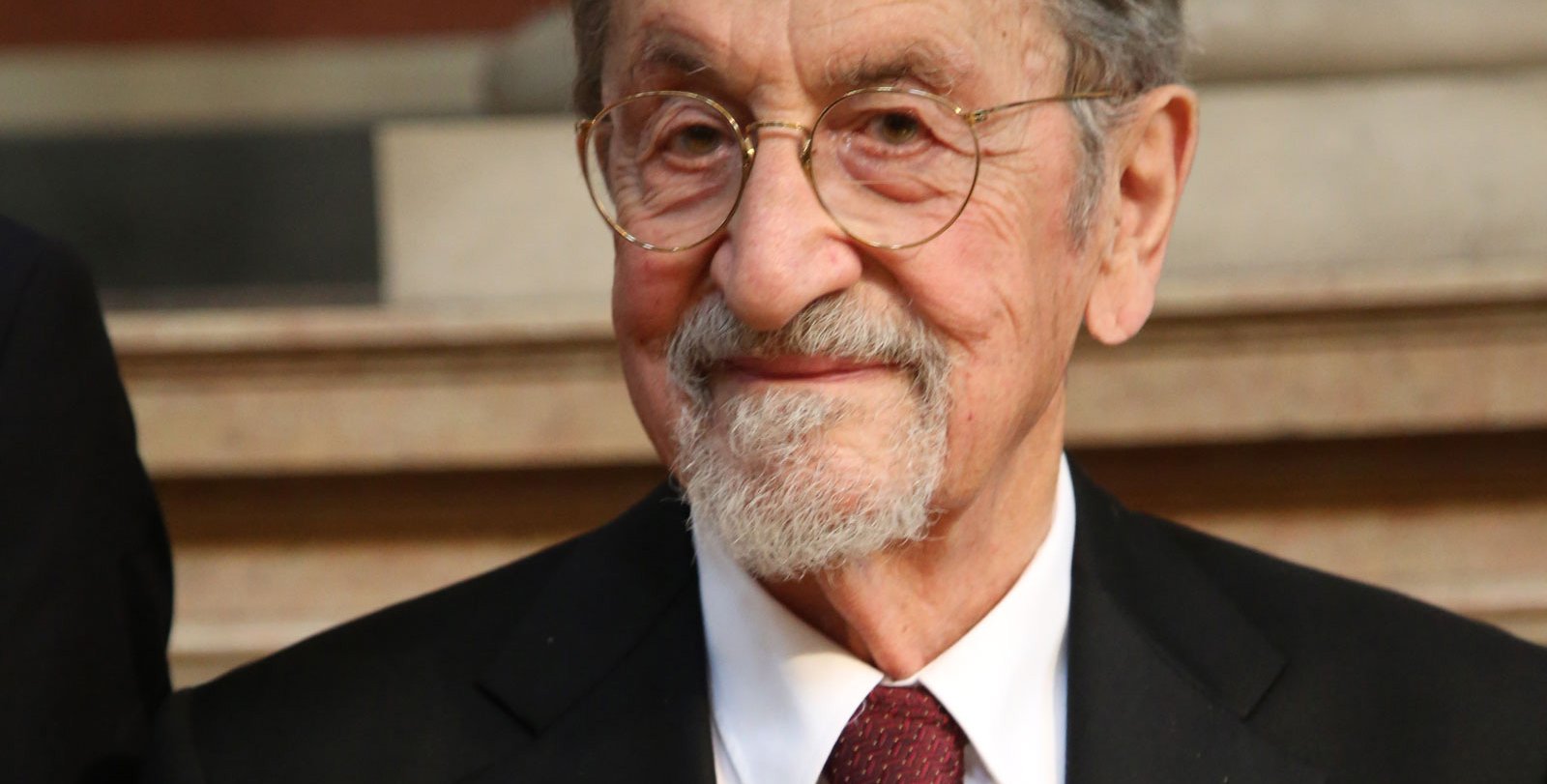 A portrait of Martin Karplus, an older man wearing a dark suit and red tie.