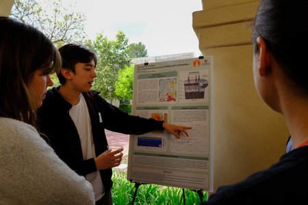 A young man wearing a white t-shirt and jacket points out something on a poster to two onlookers.