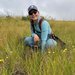 A person in blue pants and shirt with a backpack, ballcap, and sunglasses on squats amid tall grasses interspersed with yellow composite flowers and herbaceous plants, under a cloudy gray sky