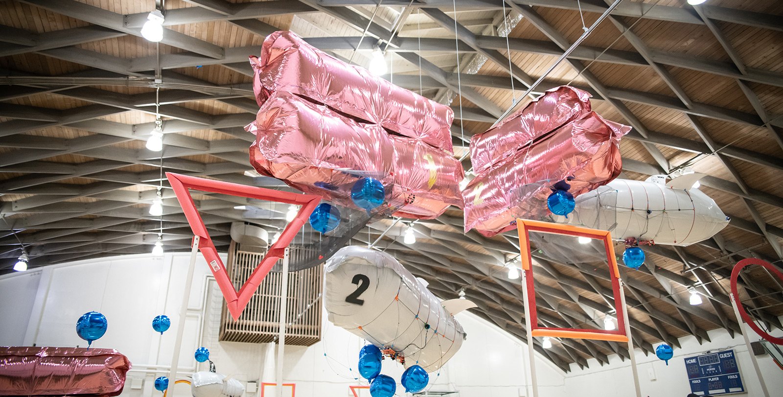 This is a picture of large metallic balloon blimps in a student competition soaring above a basketball court, trying to push blue neutrally buoyant balls through goals 10 feet up in the air.