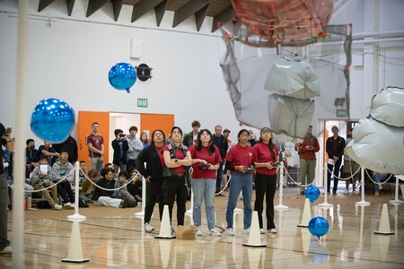 Five undergraduate students in red shirts stand in a row, looking intently at the blimps they built. Three of the students are steering the airships as they maneuver above the basketball court. Onlookers are following along in the background.