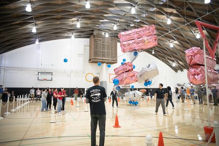 students stand around a gym with white and pink blimps flying above