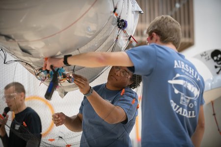 Two undergraduate students in blue shirts work with the wires underneath a white blimp.