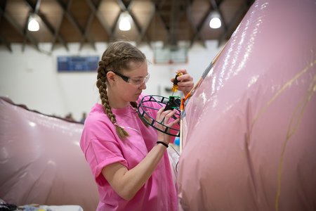 An undergraduate student in a pink shirt uses a screwdriver to make adjustments to the fan on the back of one of her team's blimps.