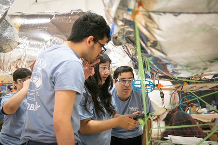 Several students in light blue shirts stare intently at the undercarriage of one of their blimps. One student holds a transmitter that will steer the silver blimp with all of the electrical components shown on the undercarriage.