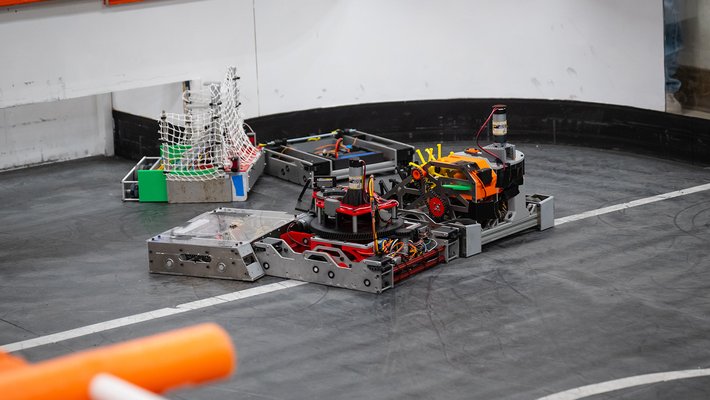 A close-up photo showing five robots battling each other, almost tangled up, on a custom-built hockey rink with white walls, an orange rim, and a black ferromagentic surface.