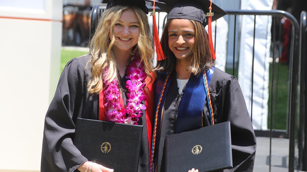 Two women in graduation robes stand next to one another holding their diplomas. The woman on the left has blond hair and a pink flowered lei; the woman on the right has shoulder-length medium brown hair and wears a blue sash.