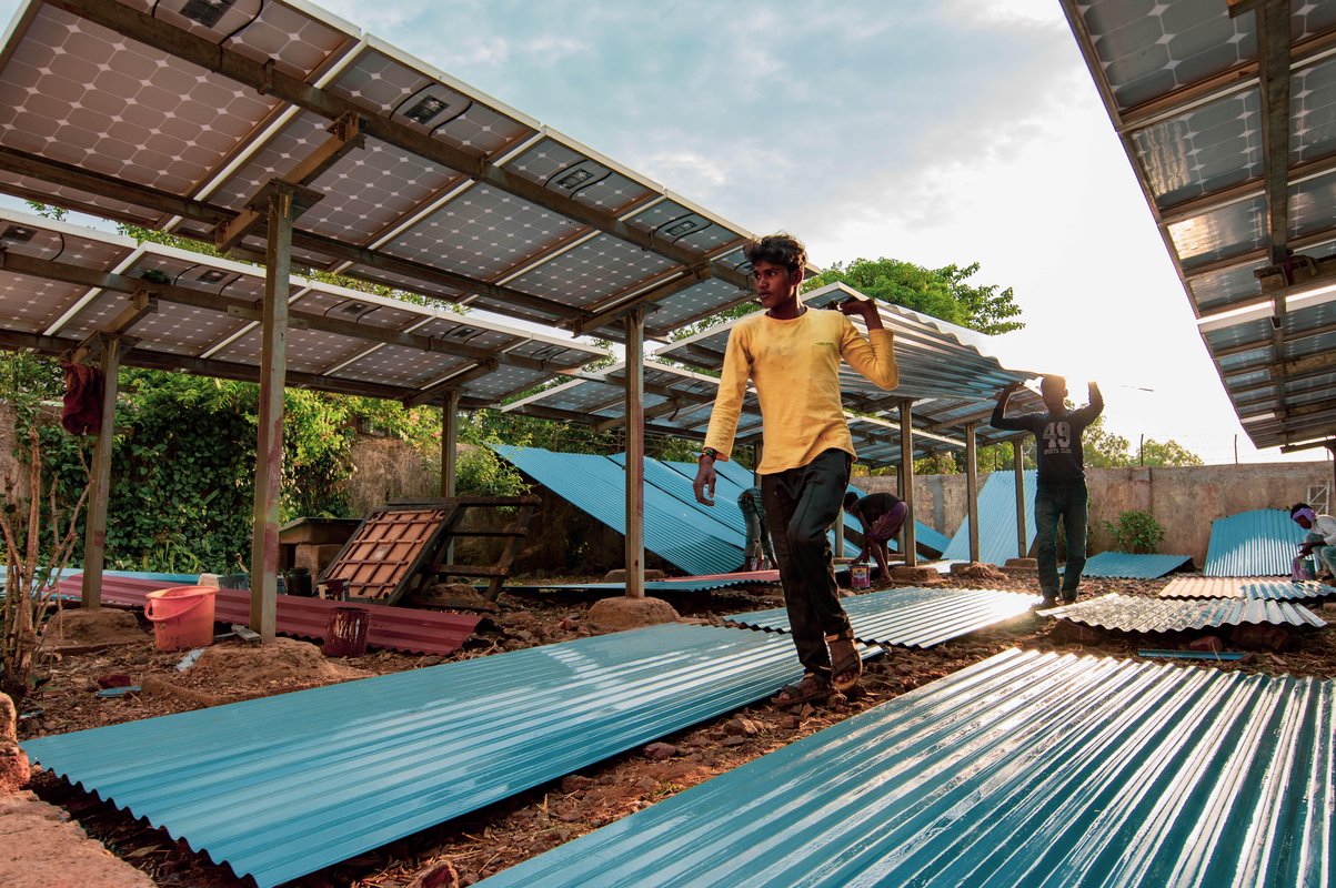 Two men carrying a roofing sheet through an area where solar panels have been set up and additional roofing material is laid out on the ground.