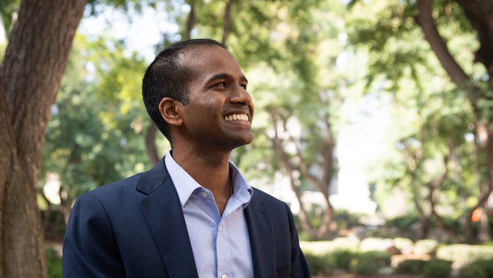 A portrait of Karthish Manthiram. He wears a button-down shirt and blazer and smiles. Trees are in the background.