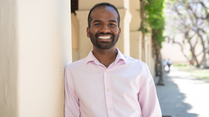 A man with brown skin and a beard wearing a pink button up shirt smiles warmly at the camera while leaning against a pillar.