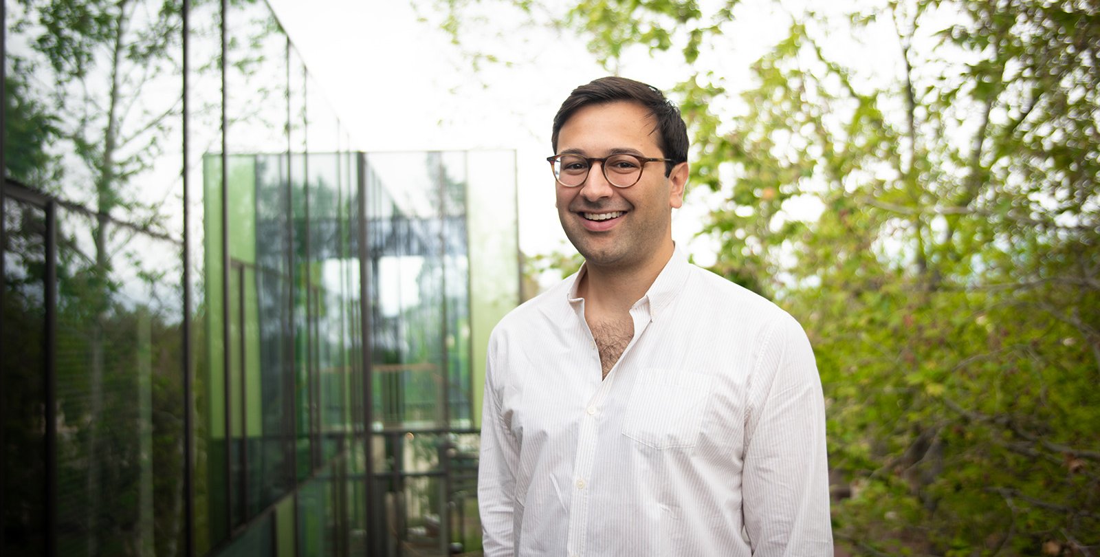 A portrait of Eric Mazumdar. He is outdoors with his hands in his pockets and smiles at the camera.