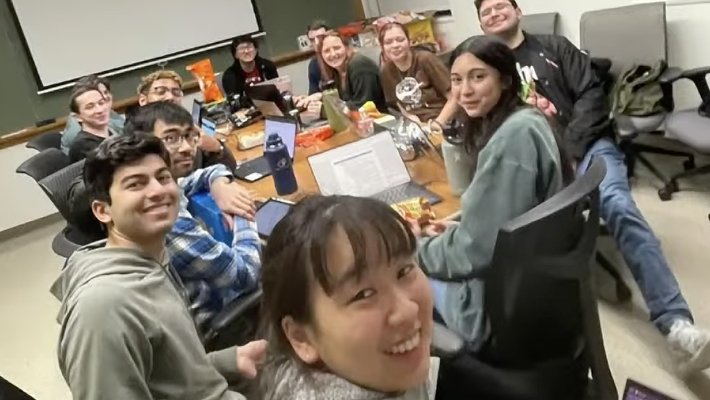 a group of undergraduate students are sitting around a table, smiling at the camera