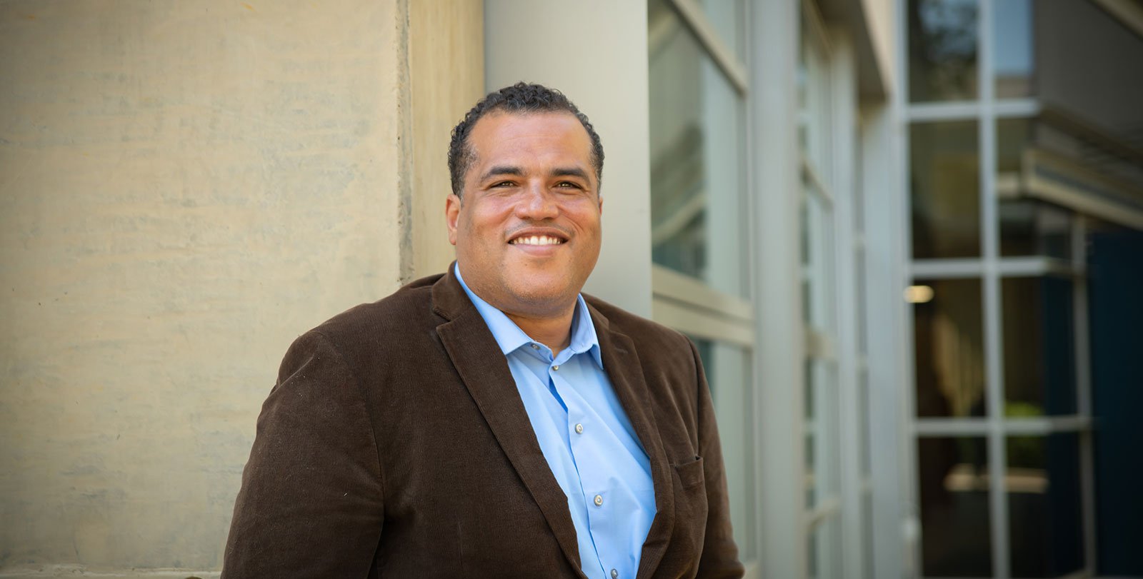 Hosea Nelson poses in front of a campus building, smiling. He wears a button-down shirt and a blazer.