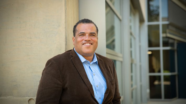 Hosea Nelson poses in front of a campus building, smiling. He wears a button-down shirt and a blazer.