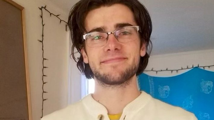 A portrait of Noah Dayton Peake. He is indoors and smiling at the camera. He wears glasses and has a short beard.