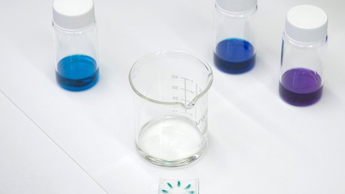 A thin piece of plastic with a colored radial pattern sits on a work bench surrounded by glass vials of brightly colored dyes.