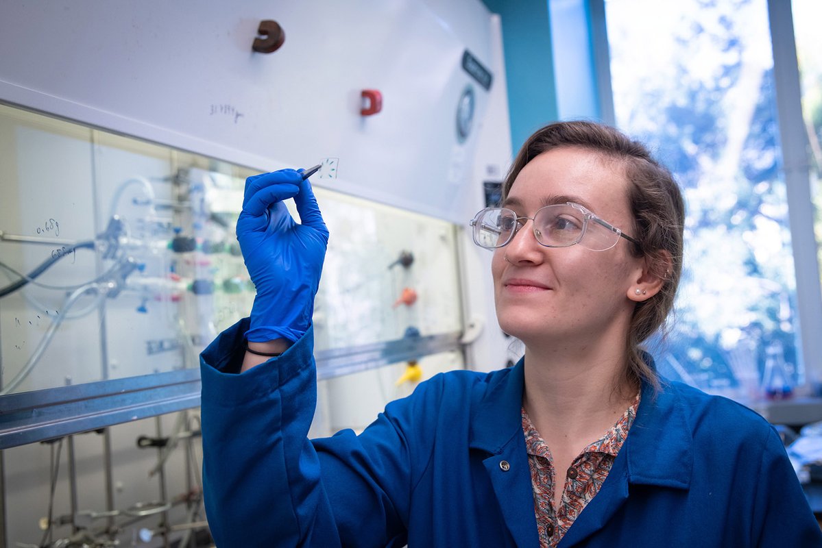 A woman holds aloft a piece of plastic with a pair of tweezers. She wears a lab coat and safety glasses. A chemistry laboratory can be seen in the background.