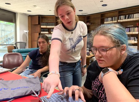 Three high school students sti at a table in a library. Two are discussing something on a computer screen.