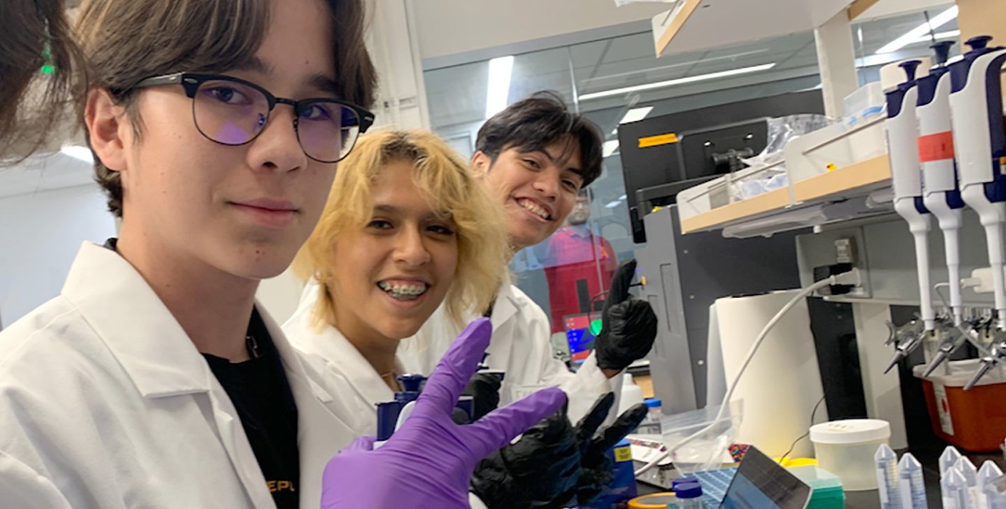 Four students are working in a scientific lab wearing rubber gloves, and using scientific instruments and a computer. All are smiling and facing the camera.