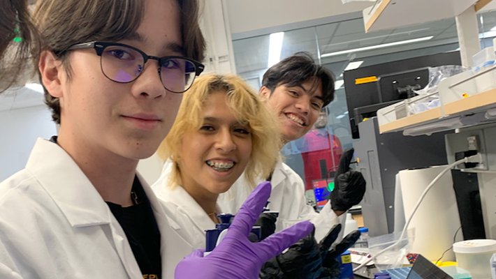Four students are working in a scientific lab wearing rubber gloves, and using scientific instruments and a computer. All are smiling and facing the camera.