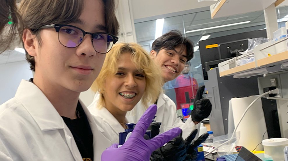 Four students are working in a scientific lab wearing rubber gloves, and using scientific instruments and a computer. All are smiling and facing the camera.