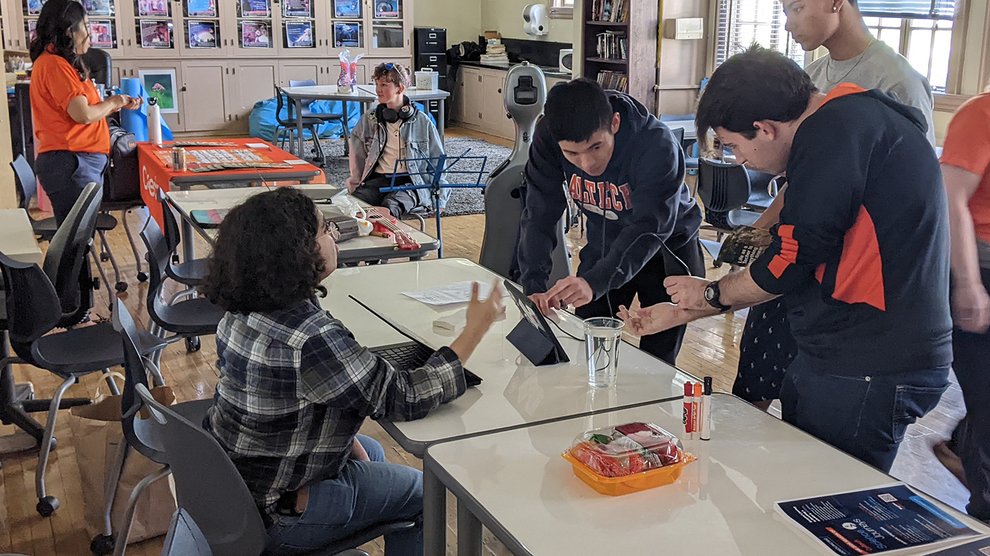 Four people gather around a table: one seated and one standing, looking on, one looking at an iPad, and another using an ultrasound imager on their arm
