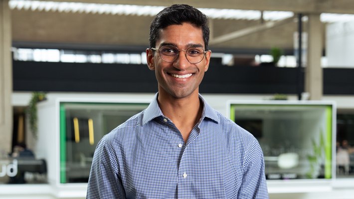 Eshaan Patheria wears round glasses and a checked blue button-up shirt and smiles at the camera. A modern urban structure is behind him.