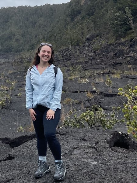 A person in black pants, a blue jacket, pink tank, and necklace stands on a dark gray ground of lava rock dotted with vigorous plants, with green trees rising on a steep hills in the background