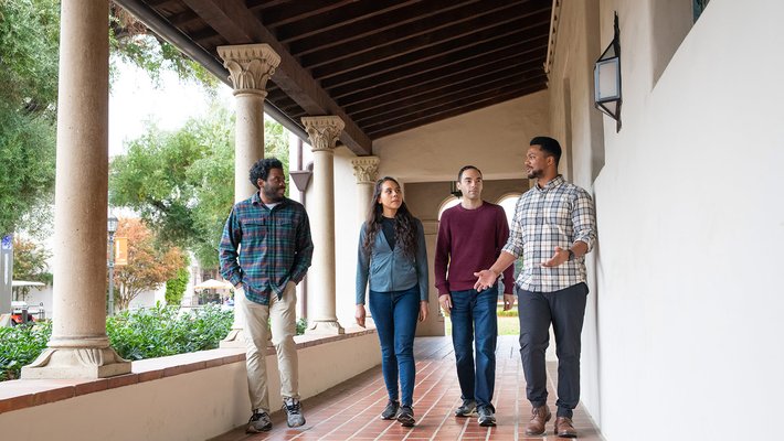 Four scholars talk amongst themselves as they walk alongside a column-lined hallway