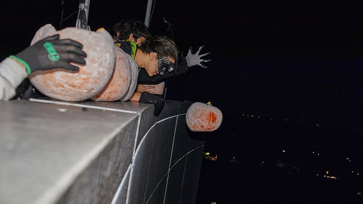 Masked woman drops a pumpkin from atop a building