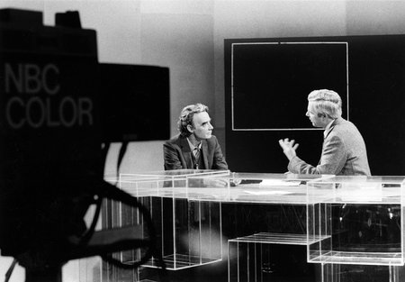 James Quirk sits and converses with an unnamed news anchor at a desk in a television studio. In the foreground, a TV camera labeled "NBC COLOR" can be seen.