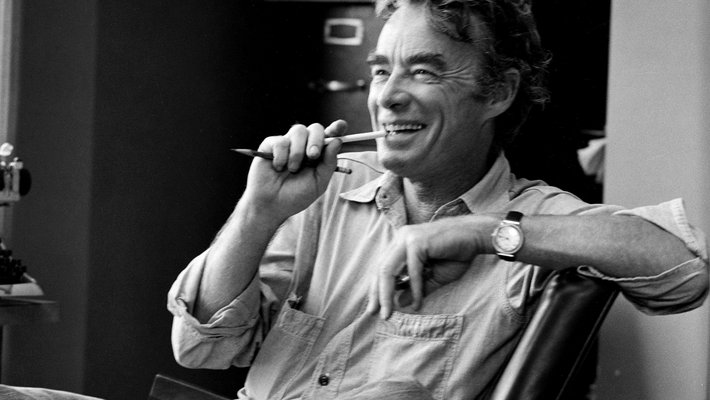 In this black-and-white photo, James Quirk sits casually in a chair, laughing and biting a pencil. He appears to be mid conversation