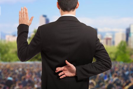 A politician stands on a stage in front of a large crowd taking an oath of office. His fingers are crossed behind his back 