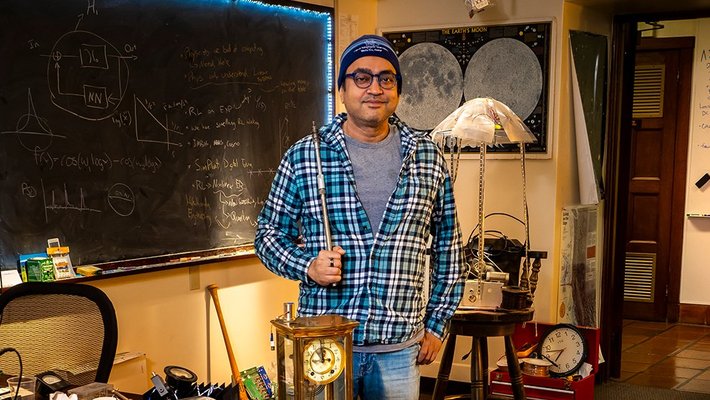 Rana Adhikari stands in his office wearing a plaid shirt and beanie, surrounded by clocks and maps and a chalkboard