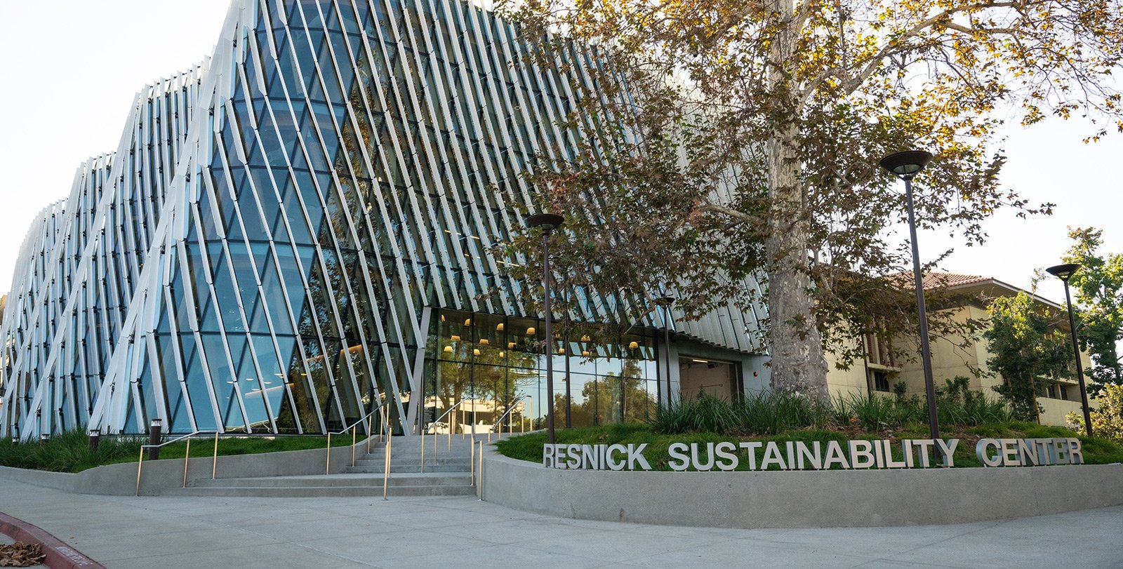 A building with a curved glass facade and diagonal shade fins on the exterior is shown with a sign reading Resnick Sustainability Center in silver letters in the foreground.
