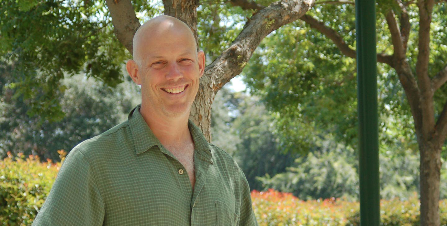 A man wears a green button down shirt and smiles for a photo outdoors