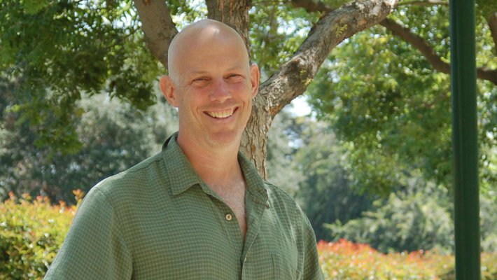 A man wears a green button down shirt and smiles for a photo outdoors