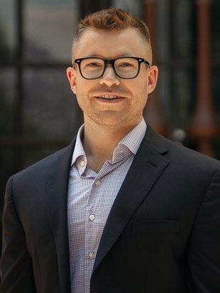 A portrait of Ryan Hadt. He wears a blazer and button-down shirt and glasses.