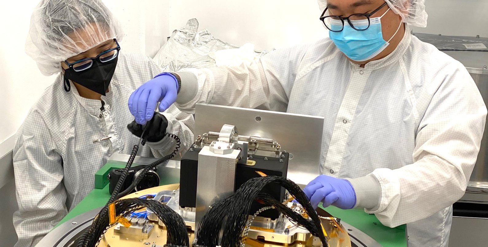 Scientists working on an instrument in lab, wearing protective suits.
