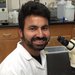 A portrait of Shashank Gandhi. He wears a lab coat and smiles while sitting in front of a microscope.