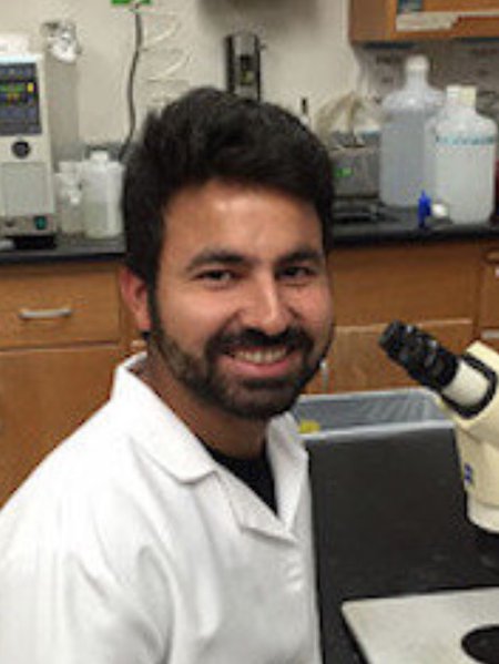 A portrait of Shashank Gandhi. He wears a lab coat and smiles while sitting in front of a microscope.