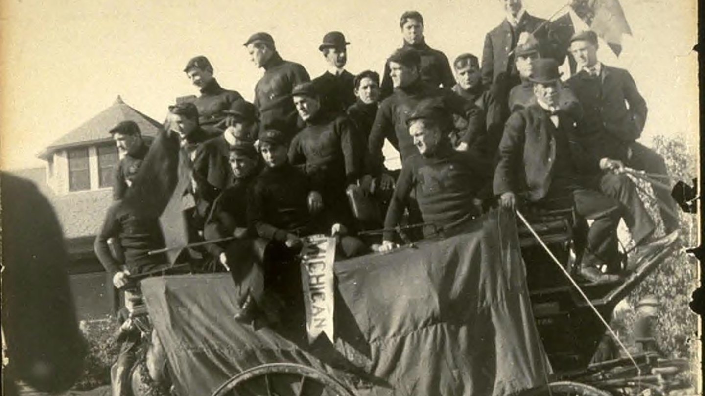 The University of Michigan football team stands crowded in a single carriage during the 1902 Rose Parade, all wearing dark Michigan sweaters in this black and white photo.