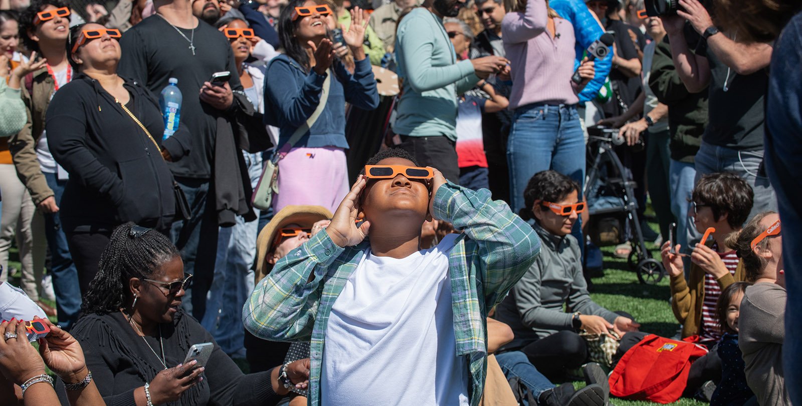 A large group of people wearing orange eclipse glasses gathered on Caltech's athletic fields. They are looking up toward the sky, observing an eclipse.