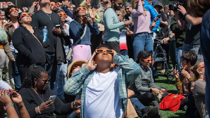 A large group of people wearing orange eclipse glasses gathered on Caltech's athletic fields. They are looking up toward the sky, observing an eclipse.