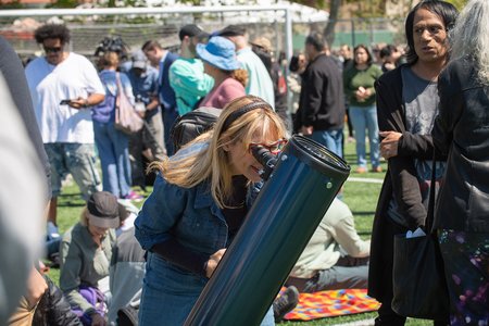 A person peers through a solar telescope, observing the eclipse amid a crowd gathered with many sitting on picnic blankets. The setting is an open field and communal atmosphere with a soccer goal visible in the background.