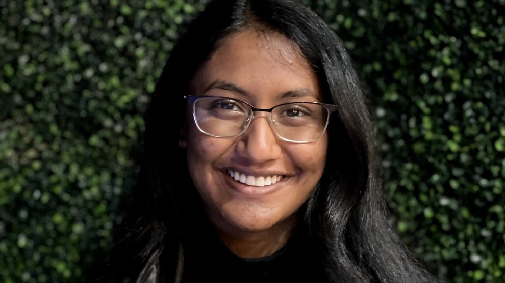 A person in a black shirt and jacket with long hair and wire-rimmed glasses smiles, standing in front of a wall of plants