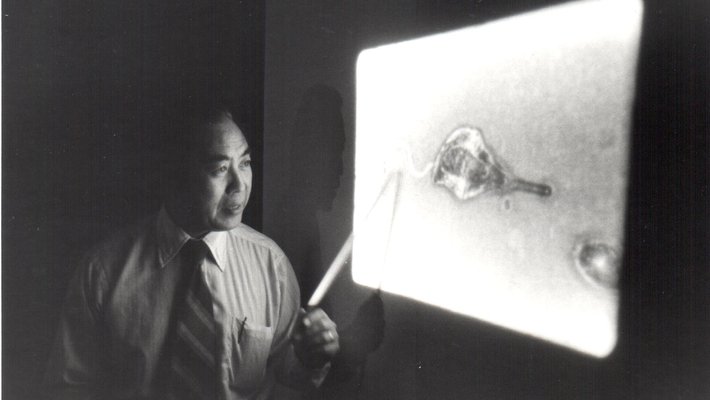 A black-and-white photo of a man wearing a shirt and tie stands at a projector screen in a darkened room. He points at the image on the screen.