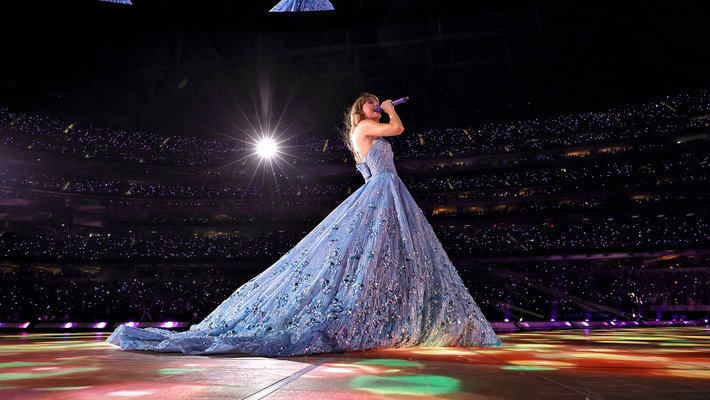 Taylor Swift, seen from the side, wearing a shimmering blue ball gown. In the background, you can see many tiny lights from the thousands of fans in SoFi Stadium.