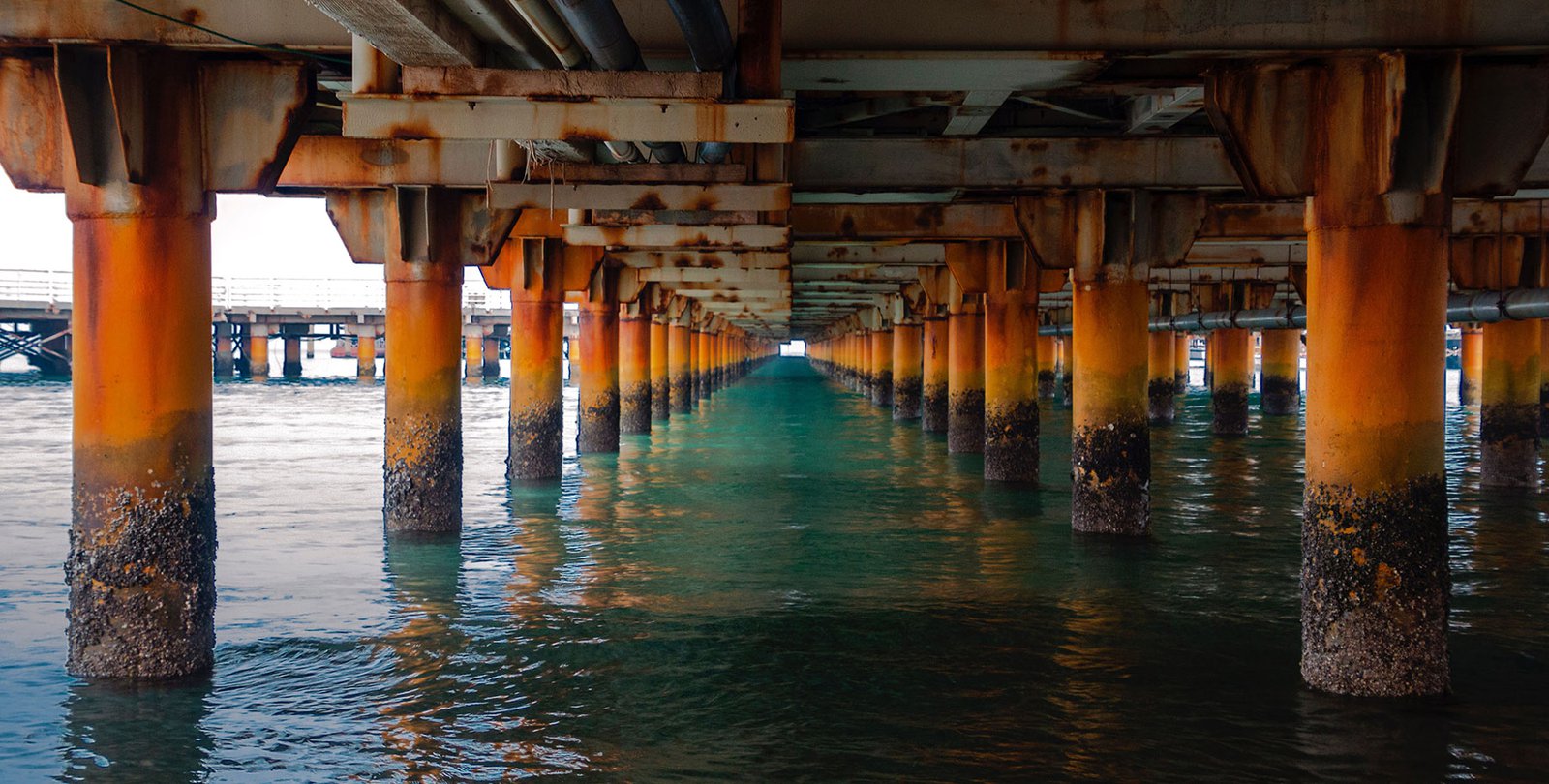 An image of the underside of a rusty bridge over a body of water.
