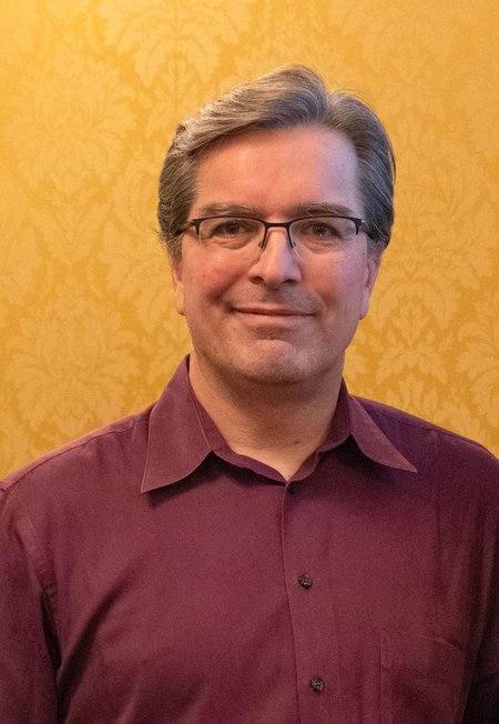 Portrait of Mike Vicic. He wears glasses and a maroon shirt and is photographed against a background of yellow wallpaper.